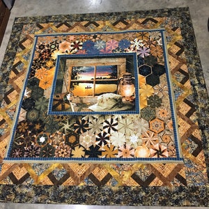 May include: A quilt with a patchwork design featuring a variety of geometric shapes in shades of brown, blue, and beige. The center of the quilt features a window scene with a lake and a boat in the distance.
