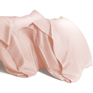 May include: Two blush pink pillowcases made of soft, smooth fabric. The pillowcases are folded and arranged on a white background.