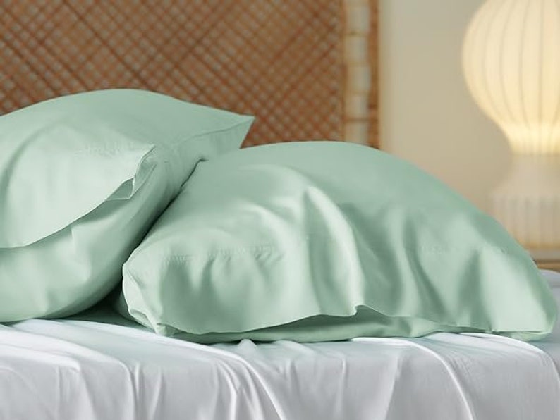 May include: Two light green pillowcases made of soft, smooth fabric. The pillowcases are lying on a white bed sheet.