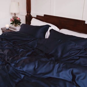 May include: A bed dressed with navy blue bedding, including a duvet and pillows. The bed has a dark wooden headboard and frame. A bedside table with a lamp, flowers, and a candle is visible.