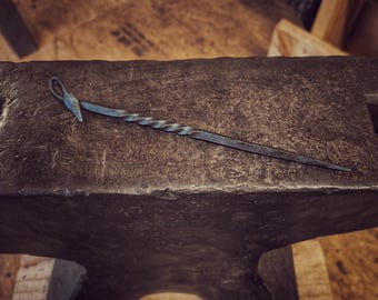 May include: A hand-forged metal skewer or pick featuring a twisted central shaft, a pointed tip, and a looped handle end with a small leaf-shaped detail. The dark, textured iron object rests horizontally on a heavy, weathered metal anvil.