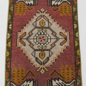 Puede incluir: Una alfombra pequeña y rectangular con un diseño turco tradicional. La alfombra tiene un fondo rojo con un medallón central blanco y beige. La alfombra tiene un borde con flecos.
