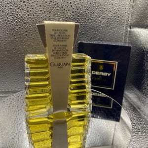 May include: A clear glass perfume bottle with a yellow liquid inside. The bottle has a unique, geometric design and a paper label with text in French. A dark blue box with gold lettering is in the background.