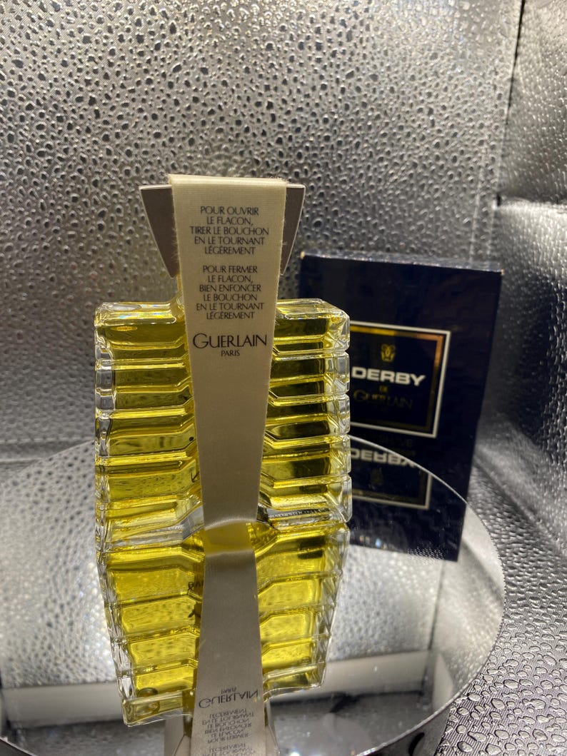 May include: A clear glass perfume bottle with a yellow liquid inside. The bottle has a unique, geometric design and a paper label with text in French. A dark blue box with gold lettering is in the background.