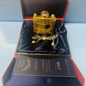 Op de afbeelding: Een gouden parfumfles met een gouden ketting en een zwart-gouden kwast. De fles zit in een rode doos met een zwarte binnenkant. De doos bevat een witte kaart met de tekst "Objet Rare OPIUM Parfum ZERTIFIKA Limitierte Auflage Jedes Objekt ist von gefertigt und zertifizik Zertifikats-Nummer 7 282 YVES SAINT LAURENT".