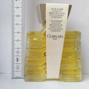 May include: A rectangular perfume bottle with a silver cap and a clear, textured body filled with yellow liquid. A white label with French text is attached. The bottle is approximately 3 inches tall, as measured by a ruler.
