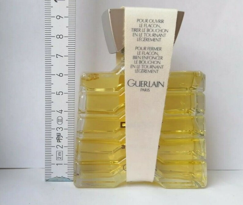 May include: A rectangular perfume bottle with a silver cap and a clear, textured body filled with yellow liquid. A white label with French text is attached. The bottle is approximately 3 inches tall, as measured by a ruler.