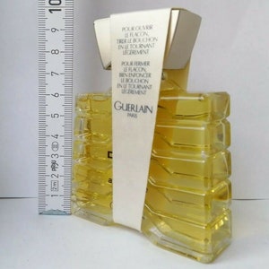 May include: A rectangular perfume bottle with a yellow liquid inside. The bottle has a unique, geometric design and is wrapped with a white label that has French text. The label reads "GUERLAIN PARIS". A ruler is placed next to the bottle.