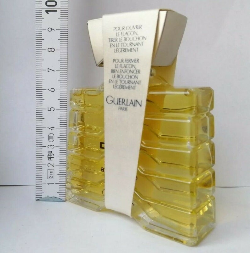 May include: A rectangular perfume bottle with a yellow liquid inside. The bottle has a unique, geometric design and is wrapped with a white label that has French text. The label reads "GUERLAIN PARIS". A ruler is placed next to the bottle.