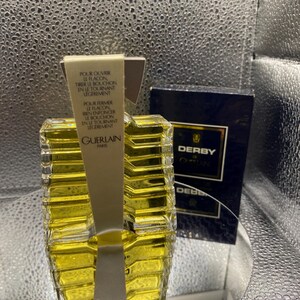 May include: A clear glass perfume bottle with a yellow liquid inside, labeled "GUERLAIN PARIS". The bottle has a unique geometric design and is accompanied by a dark blue box labeled "DERBY". Instructions are printed on a white label.