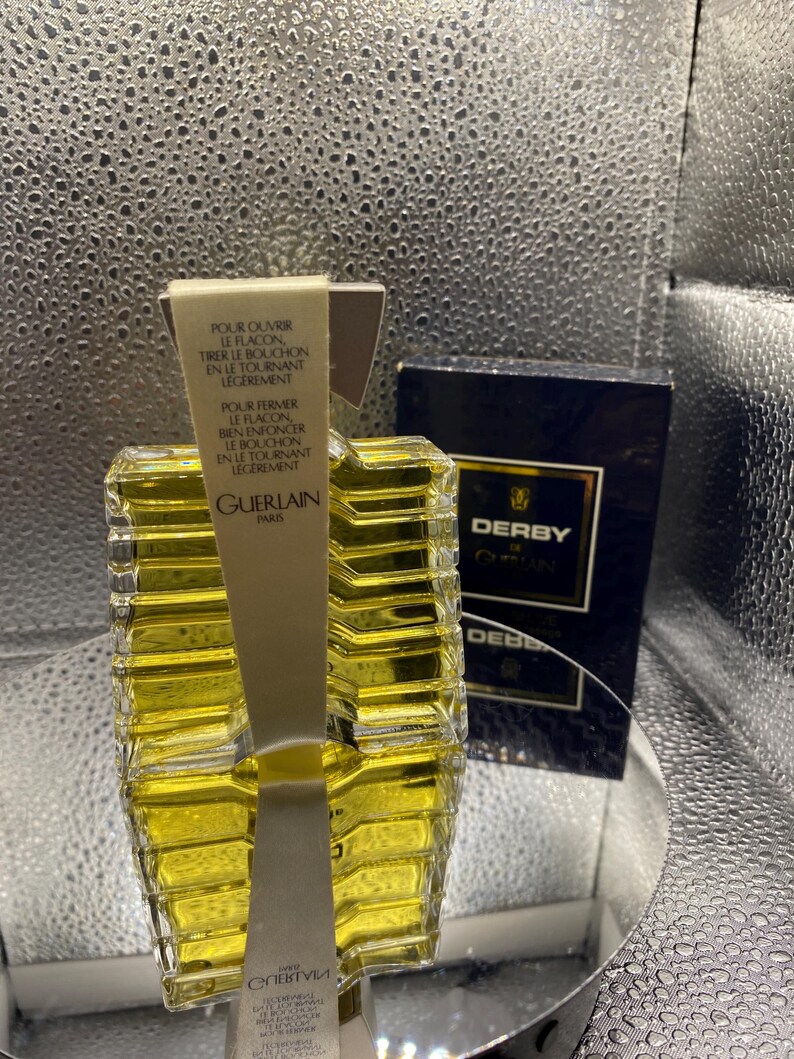 May include: A clear glass perfume bottle with a yellow liquid inside, labeled "GUERLAIN PARIS". The bottle has a unique geometric design and is accompanied by a dark blue box labeled "DERBY". Instructions are printed on a white label.