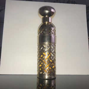 Könnte beinhalten: Eine goldfarbene Parfümflasche mit einem dekorativen, durchbrochenen Design. Die Flasche hat einen abgerundeten Deckel und einen zylindrischen Körper mit einem geometrischen Muster. Der Markenname "Guerlain" ist auf der Basis sichtbar.
