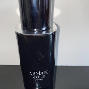 May include: A black Armani Code Parfum bottle with a silver rim and a black cap. The bottle has the Armani Code logo in white. The bottle is on a reflective surface.