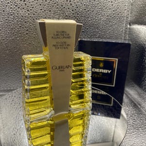 May include: A Guerlain perfume bottle with a rectangular design, filled with a yellow liquid. The bottle has a paper label with instructions and the Guerlain Paris logo. A dark blue box with the word "DERBY" is visible in the background.