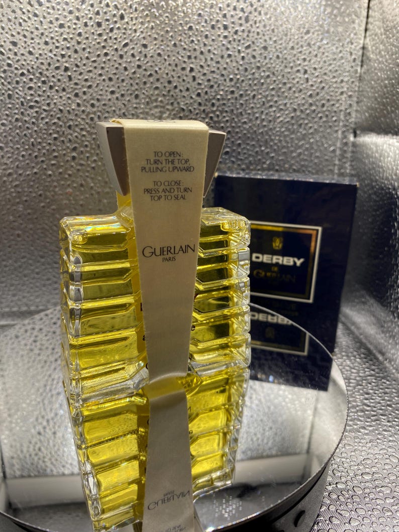 May include: A Guerlain perfume bottle with a rectangular design, filled with a yellow liquid. The bottle has a paper label with instructions and the Guerlain Paris logo. A dark blue box with the word "DERBY" is visible in the background.