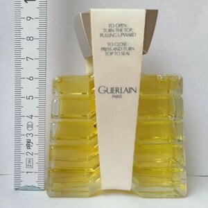 May include: A rectangular perfume bottle with a silver cap and a cream-colored label. The bottle contains a yellow liquid and has the text "GUERLAIN PARIS". The label reads "TO OPEN: TURN THE TOP, PULLING UPWARD. TO CLOSE: PRESS AND TURN TOP TO SEAL."
