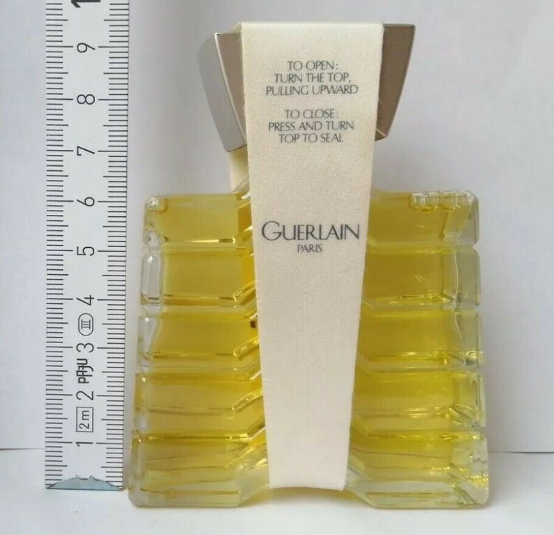 May include: A rectangular perfume bottle with a silver cap and a cream-colored label. The bottle contains a yellow liquid and has the text "GUERLAIN PARIS". The label reads "TO OPEN: TURN THE TOP, PULLING UPWARD. TO CLOSE: PRESS AND TURN TOP TO SEAL."