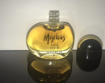 Op de afbeelding: Een heldere, ronde parfumfles gevuld met gouden vloeistof, met het label "Moschus Love Fever Perfume Oil". De fles heeft een zilveren dop en staat naast een zwarte, afgeronde deksel met een gouden band. De weerspiegeling van de fles is zichtbaar.