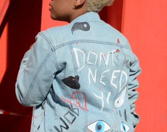 Puede incluir: Una chaqueta vaquera azul claro con detalles pintados en blanco. La parte posterior de la chaqueta tiene las palabras "Don't Need You" escritas en pintura blanca. Hay otros diseños pintados en la chaqueta, incluyendo un símbolo yin yang, un corazón y un ojo.