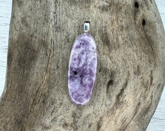 May include: A lavender-colored, oval-shaped pendant with a silver-colored bail. The pendant has a marbled appearance with darker purple and white inclusions. The pendant is displayed on a piece of weathered driftwood.