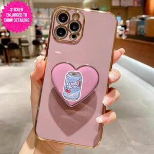 May include: A pink phone case with a gold-colored border. A pink heart-shaped phone grip with a "Girly Pop" soda can design is attached to the back of the phone case.