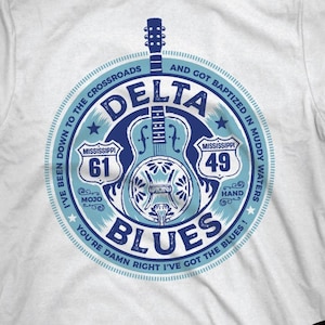 Puede incluir: Camiseta blanca con un diseño circular azul que presenta una guitarra y las palabras "Delta Blues". El diseño incluye texto que hace referencia a las carreteras 61 y 49 de Mississippi. Un logotipo de "Mojo Hand" está a la derecha.