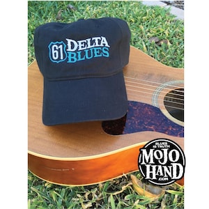 May include: Black baseball cap with the text "61 Delta Blues" in white and turquoise. The cap rests on a wooden acoustic guitar. The Mojo Hand logo is visible in the lower right corner.