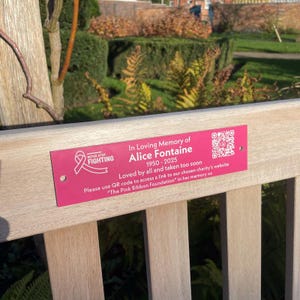 May include: A pink memorial plaque fixed to a wooden bench. The plaque reads "In Loving Memory of Alice Fontaine 1950-2025" with a pink ribbon logo and the words "Never Stop Fighting". A QR code is also present.