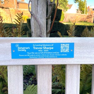 May include: A blue plaque on a wooden bench, commemorating Trevor Sharpe (1952-2025). The plaque, with text about the Alzheimer's Society, includes a QR code. The background features a tree and foliage, suggesting an outdoor setting.