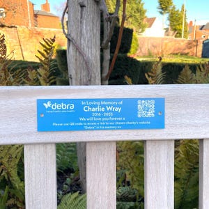 May include: A blue plaque on a wooden bench, commemorating Charlie Wray (2016-2025). The plaque features the "debra" charity logo and a QR code. The bench is set amongst foliage. The text reads "We will love you forever x".