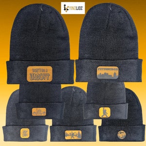 May include: Nine black knit beanies with different embroidered patches. The patches feature various designs, including a city skyline, a monster, a cartoon character, and the words "Don't be a Jagoff".
