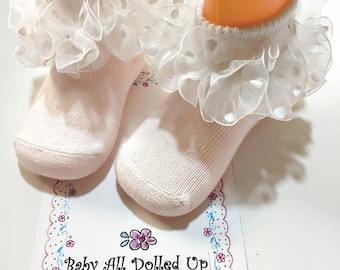 May include: A pair of white baby socks with a white lace ruffle trim and small white polka dots. The socks are on a white background with a pink floral design and the text "Baby All Dolled Up".