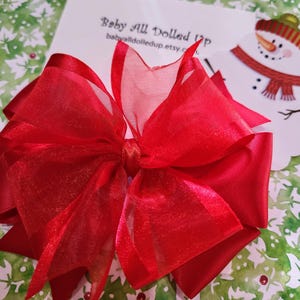 May include: A large red sparkly bow made of sheer fabric. The bow is tied in a knot in the center and has a layered look.