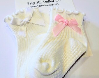 May include: A pair of white baby socks with a ruffled top and a small bow. One sock has a white bow, and the other has a pink bow. The soles have a non-slip grip. The text "Baby All Dolled Up" is visible.