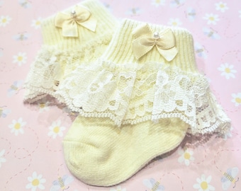 May include: A pair of yellow baby socks with white lace trim and a satin bow with a pearl accent.
