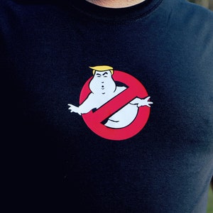Puede incluir: Una camiseta negra con un círculo rojo y una figura de fantasma blanca en su interior. El fantasma tiene el pelo amarillo y una expresión seria. El círculo tiene una línea diagonal que lo atraviesa.