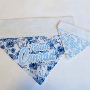 May include: Two dog bandanas. One large bandana with blue floral pattern and the text "Team Conrad" in light blue. The second bandana is smaller, with a similar floral pattern and the text "Team Jeremiah".