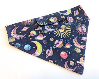 Puede incluir: Un pañuelo negro con un estampado celestial que presenta lunas, estrellas y planetas en tonos de rosa, amarillo y azul. El pañuelo tiene un pequeño bolsillo con cierre de botón.