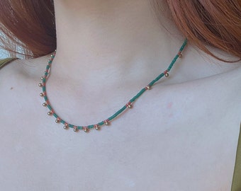 May include: A green and red beaded necklace with gold accents. The necklace is worn by a person with long red hair.