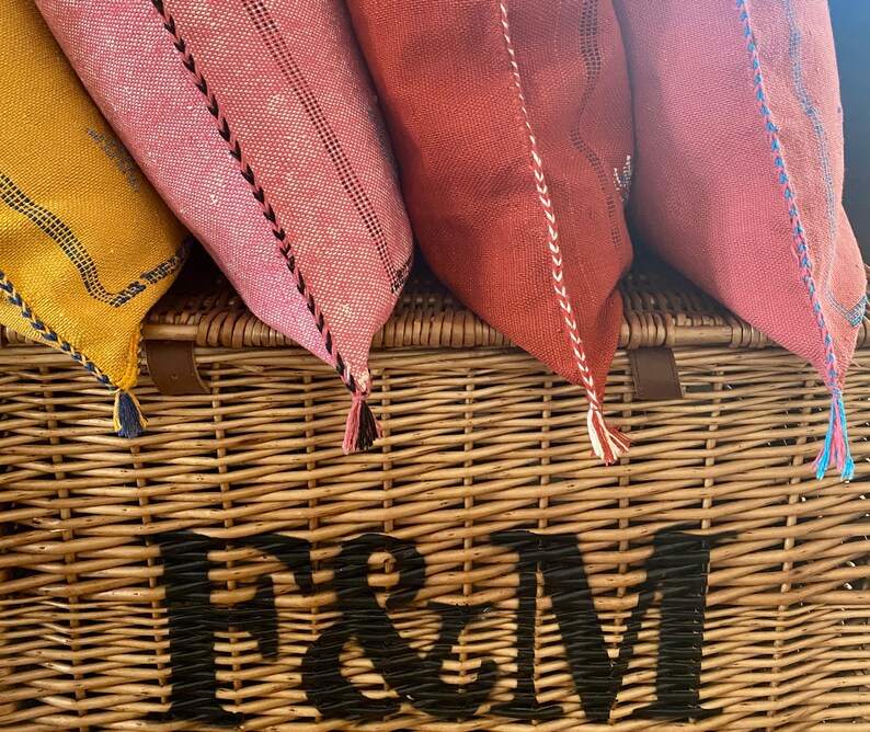 May include: Four colorful throw pillows with tassels are arranged on top of a wicker basket with the text "Fortnum & Mason" printed on the front.