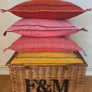May include: Four colorful throw pillows stacked on top of a wicker basket with the letters "F&M" printed on the front.