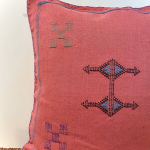 May include: A pink throw pillow with blue and black embroidered geometric patterns. The pillow has a fringe detail on one corner.