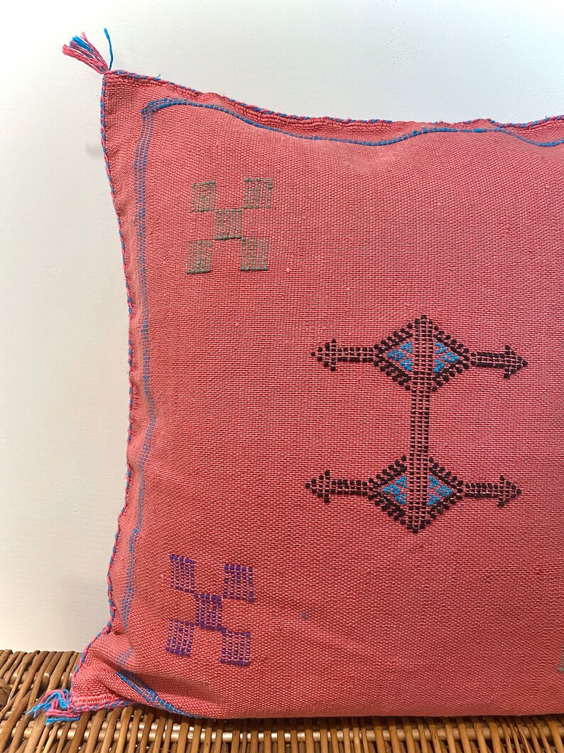 May include: A pink throw pillow with blue and black embroidered geometric patterns. The pillow has a fringe detail on one corner.
