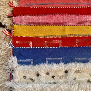 May include: A stack of colorful woven fabric squares with fringe edges. The top square is white with silver sequins. The squares below are red, blue, yellow, red, pink, and red.