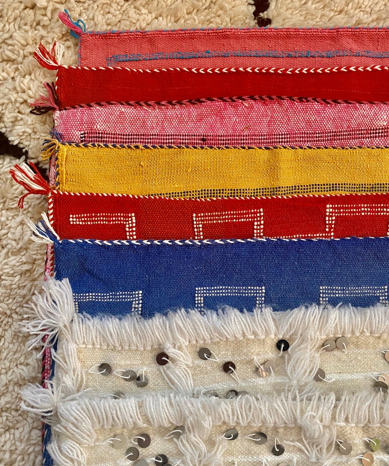May include: A stack of colorful woven fabric squares with fringe edges. The top square is white with silver sequins. The squares below are red, blue, yellow, red, pink, and red.