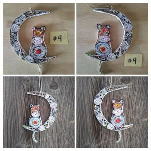 May include: A handmade paper mache cat sitting on a crescent moon. The cat and moon are decorated with colorful paper scraps. The cat is white with black and red accents. The moon is white with black and red accents. The cat and moon are hanging from a string. The image includes the text "#4".