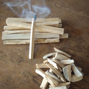 May include: A lit palo santo stick with blue smoke rising, next to a pile of unlit sticks and wood shavings. The wood is light brown, and the background is a dark brown wooden surface.