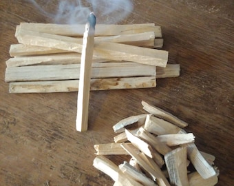 May include: A lit palo santo stick with blue smoke rising, next to a pile of unlit sticks and wood shavings. The wood is light brown, and the background is a dark brown wooden surface.