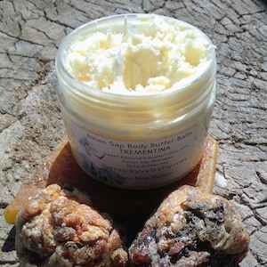 May include: A clear jar filled with a light yellow body butter balm. The label reads "Piñon Sap Body Butter Balm TREMENTINA". The jar sits on a piece of amber-colored resin, with two pieces of resin in the foreground.