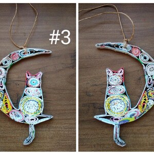 May include: A colorful paper mache cat sitting on a crescent moon. The cat and moon are made of rolled up paper and have a multi-colored pattern. The cat is white with red, blue, and yellow accents. The moon is white with green, yellow, red, and blue accents. #3